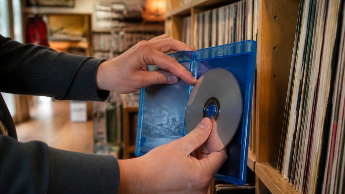 Hands placing a Blu-ray disc into its case on a shelf of physical media in a cozy store.