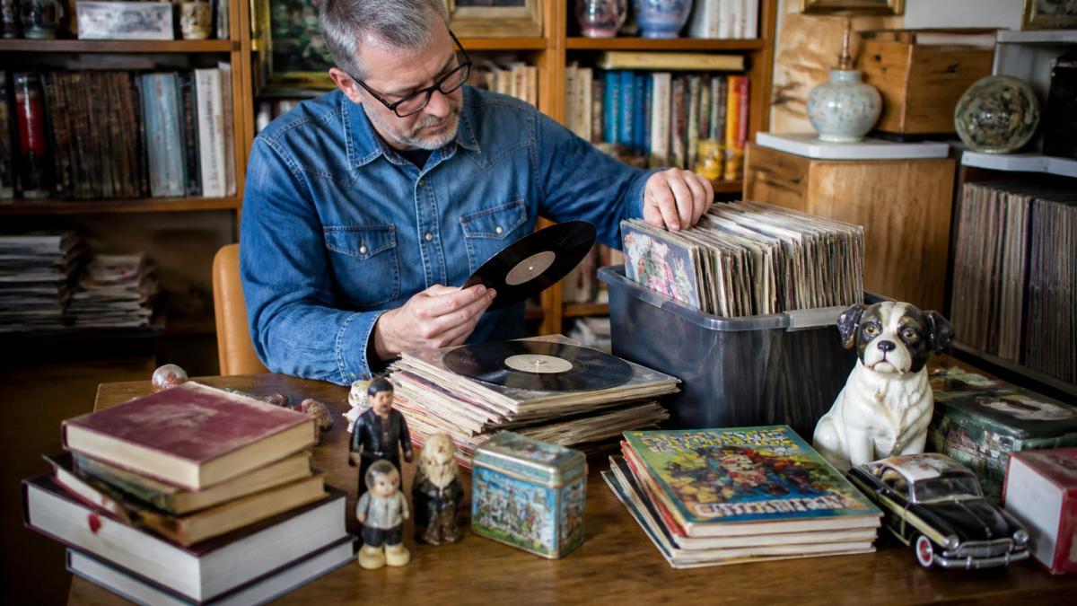 Person carefully examining and organizing vintage vinyl records, books, and collectibles on a wooden table with a notebook and valuation notes nearby