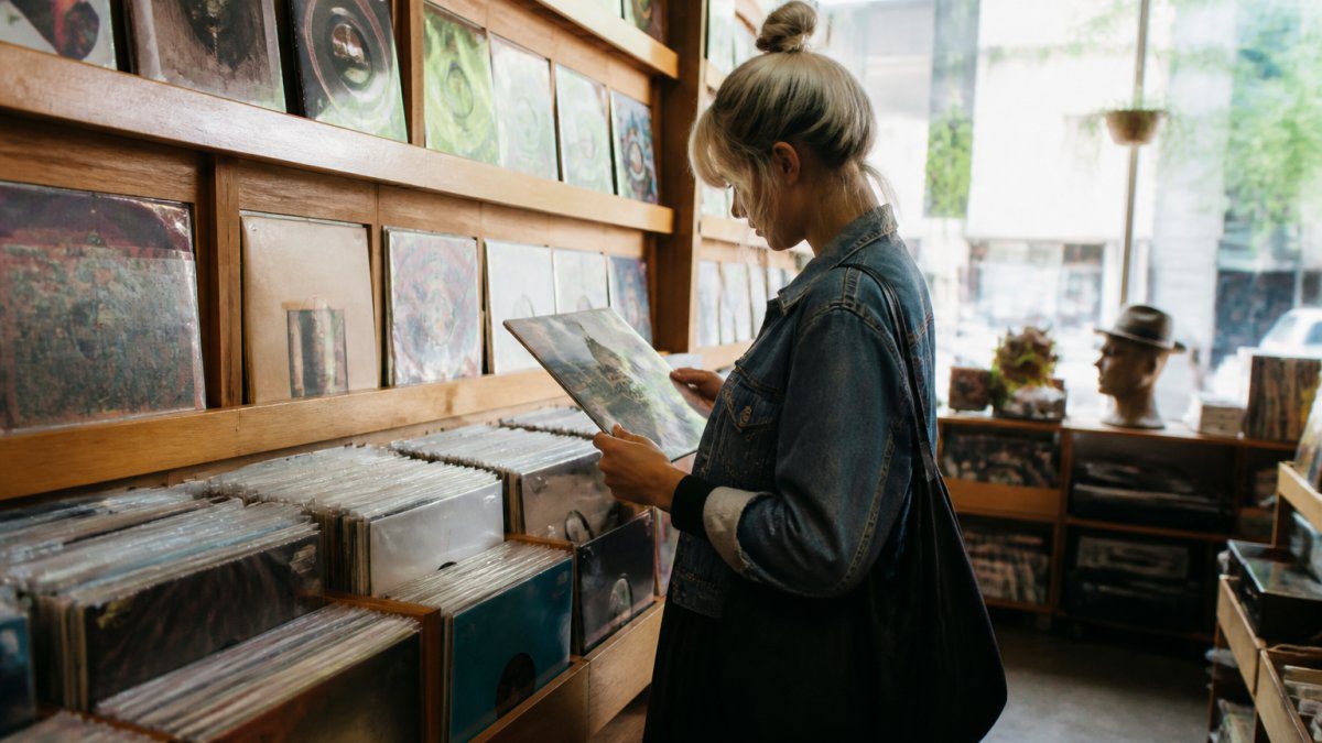 Person browsing vinyl records on display shelves in a record store, carefully examining album covers with focused attention and natural light filtering through storefront windows